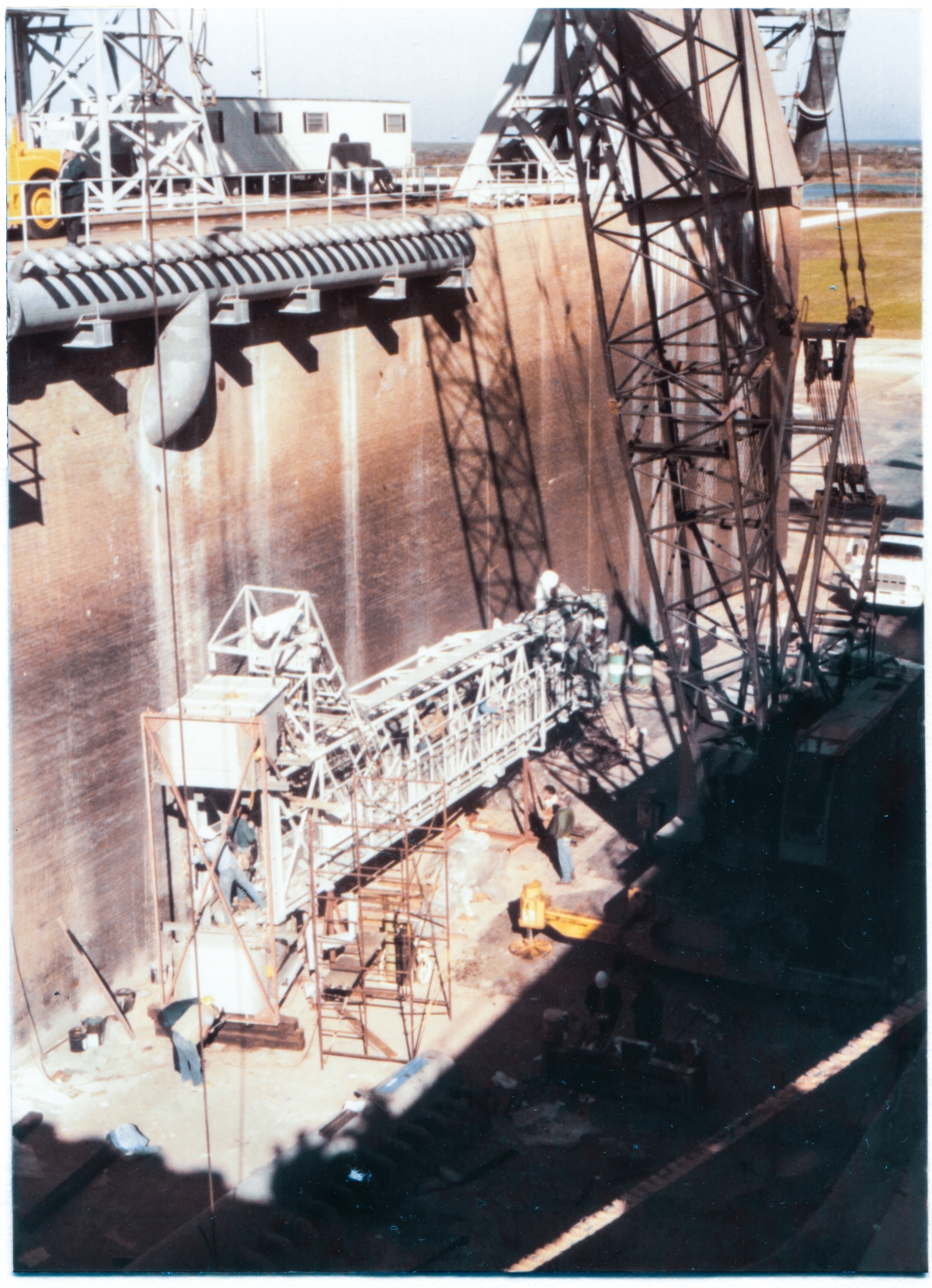 Image 129. At Space Shuttle Launch Complex 39-B, Kennedy Space Center, Florida, preparatory work on the Orbiter Access Arm (OAA) is in progress, prior to it being hung on the tower. The OAA is sitting on metal support stanchions, down in the bottom of the Flame Trench, north of the Flame Deflector. This is the same location that work on the GOX Arm was done, prior to it getting lifted. From here, it's pretty straightforward crane work to pick it up and place it on the Pad Deck, where the final preparations will be completed, immediately prior to lifting the Arm, and hanging it on Side 1 of the FSS at elevation 200'-0”. At the time this photograph was taken, most of the work being done was Electrical and Mechanical trades work, but not every last bit of it. The Environmental Chamber (also known as the ”White Room”) has yet to be attached to the far end of the arm, opposite along its length, away from the Hinges, which attach directly to the Strongback on the FSS, and which can be seen in the lower left portion of this image. Photo by James MacLaren.